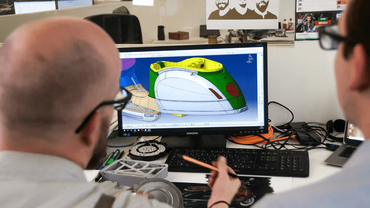 Two engineers reviewing a 3D CAD model of a mechanical part on a computer monitor.